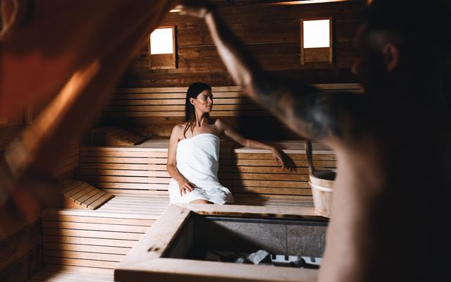 ein mann macht einen aufguss in der finnischen sauna, während die frau auf der bank sitzt und den ausblick in den garten im vulkanlandhotel legenstein genießt